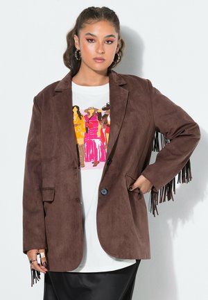 Jeune femme aux cheveux tressés porte une veste en daim marron à franges sur un t-shirt blanc imprimé et une jupe noire, posant contre un mur blanc.