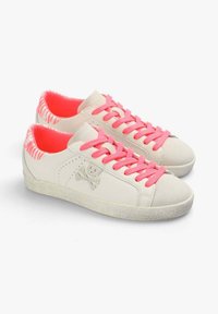 Baskets blanches en matériau lisse avec des lacets et une doublure intérieure rose vif. Elles possèdent une semelle texturée et un motif de tête de mort avec des os croisés.