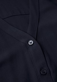 Navyblauwe, gestructureerde stoffen shirt met een V-hals en twee knopen, voorzien van een zilveren logo-knoop en een glanzende afwerking.