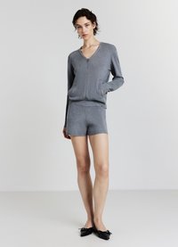 Frau in grauem Reißverschluss-Kardigan und passenden Shorts mit schwarzen Ballerinas, die vor einem schlichten hellen Hintergrund steht.