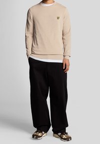 Beige knitted sweater with round neck and embroidered yellow logo on chest, paired with black loose-fitting pants and camouflage sneakers.