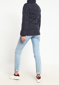 Marineblå pufferjakke med høj krave, quiltet design og slank pasform, parret med lyseblå jeans og hvide sneakers med burgunder detaljer.