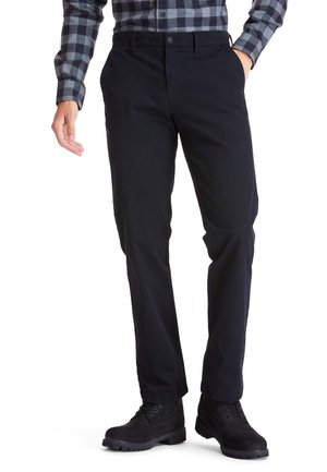 Homme portant un pantalon noir droit, des bottes noires à lacets, et une chemise à manches longues à carreaux gris et noir, avec une main dans la poche.