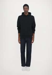 Black hooded sweatshirt with embossed logo, paired with dark blue denim jeans and white sneakers. Model stands against a plain white background.