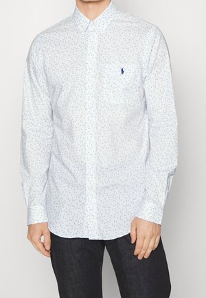 Man draagt een wit overhemd met knopen en een lichtblauw bloemenpatroon en een donkere broek, staande tegen een effen lichte achtergrond.