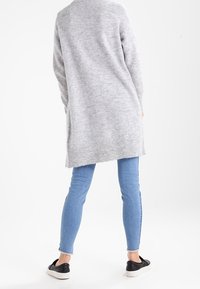Grå lång kofta i mjukt material, kombinerad med ljusblå tajta jeans och svarta sneakers. Koftan har en avslappnad passform och inga dekorationer.