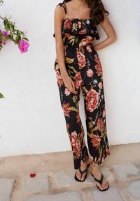 Femme portant une combinaison noire sans manches à motif floral et des sandales noires, debout contre un mur blanc avec des fleurs roses à proximité.