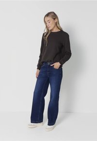 Dunkelbrauner Pullover mit rundem Ausschnitt, kombiniert mit weiten blauen Jeans. Die Jeans haben einen mittleren Waschgang und sind mit beigen Sneakers gestylt.