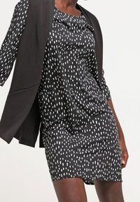 Robe noire avec un motif de pois vertical blancs ; tissu doux, longueur courte et coupe décontractée. Associée à un blazer noir ajusté.