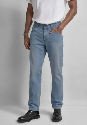 Uomo con jeans azzurri, t-shirt bianca e scarpe eleganti in pelle nera, in piedi con una mano in tasca davanti a uno sfondo neutro.