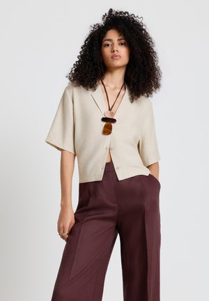 Femme aux cheveux bouclés portant une chemise beige à manches courtes boutonnée, un pantalon large bordeaux et un collier superposé avec pendentifs.