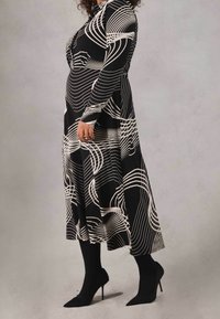 Persona incinta in piedi di profilo che indossa un vestito lungo con stampa astratta bianco e nero, collant neri e scarpe con tacco nero.