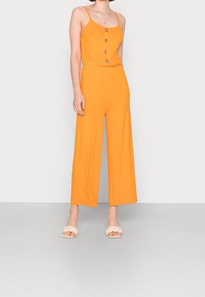 Kvinde iført en ærmeløs orange jumpsuit med vide ben og knapper foran, stående på en ensfarvet lys baggrund med hvide sandaler.