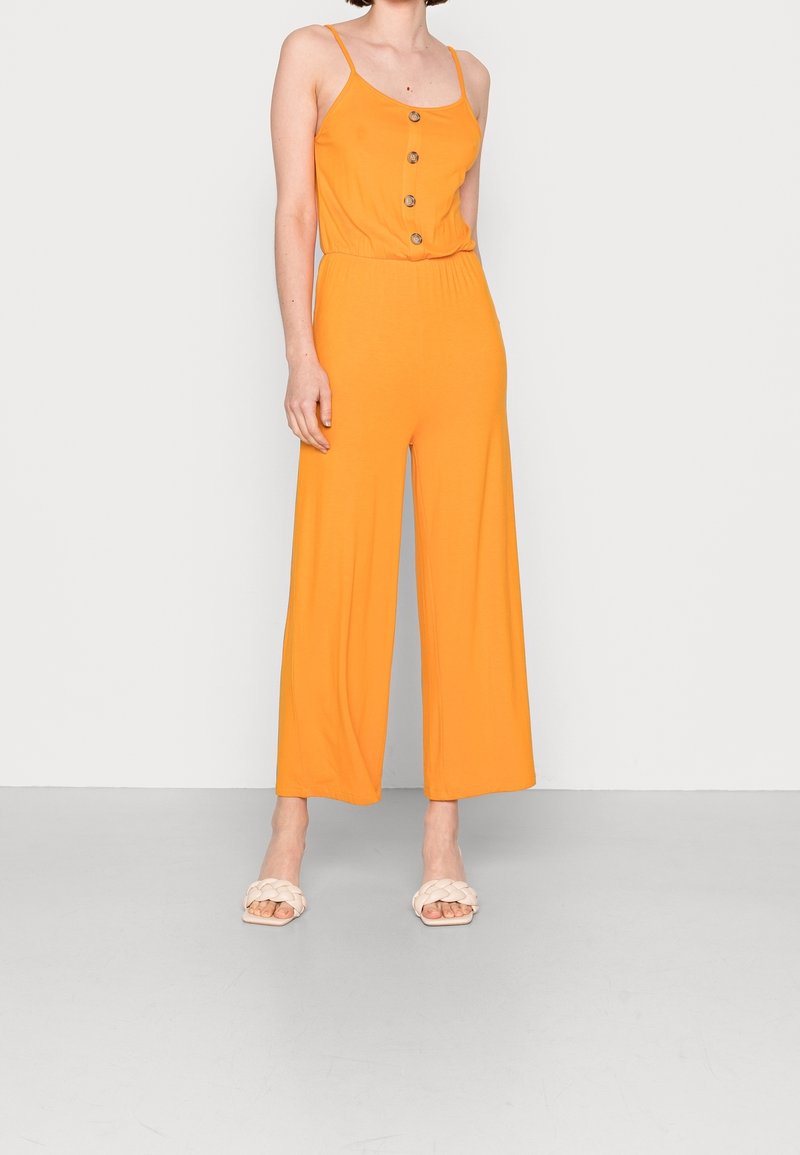 Femme portant une combinaison orange sans manches à jambes larges et boutons sur le devant, se tenant sur un fond clair uni avec des sandales blanches.