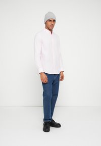Camicia rosa chiaro con bottoni e maniche lunghe, abbinata a jeans blu a gamba dritta e scarpe nere. La persona indossa un berretto grigio di maglia.