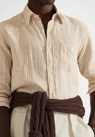 Άνδρας που φοράει πουκάμισο με μπεζ και λευκές ρίγες και κουμπιά, κρεμ παντελόνι και σκούρο καφέ πουλόβερ δεμένο γύρω από τη μέση.