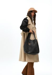 Frau in einem beigen Trenchcoat mit schwarzen Ärmeln, die eine beige Mütze trägt und eine schwarze gesteppte Handtasche hält. Sie steht vor einem weißen Hintergrund.