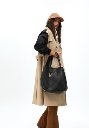 Frau in einem beigen Trenchcoat mit schwarzen Ärmeln, die eine beige Mütze trägt und eine schwarze gesteppte Handtasche hält. Sie steht vor einem weißen Hintergrund.