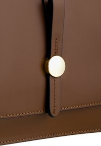 Borsa in pelle marrone con una superficie liscia, dotata di una tracolla nera e un dettaglio in hardware circolare dorato sulla parte anteriore. I bordi cuciti sono visibili.