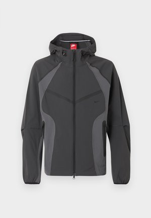 Dunkelgraue Nike Kapuzenjacke mit durchgehendem Frontreißverschluss, seitlichen Reißverschlusstaschen und elastischen Bündchen, dargestellt vor weißem Hintergrund.