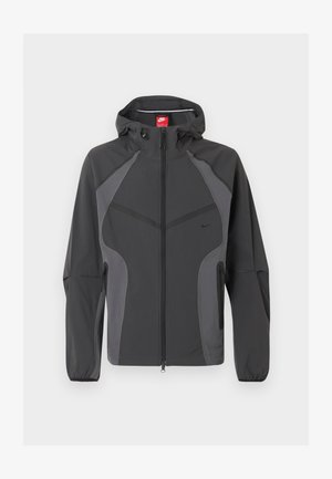 Temno siva Nike jakna s kapuco, z zadrgo po celotni sprednji strani, žepki na straneh z zadrgo in elastičnimi manšetami, prikazana na belem ozadju.