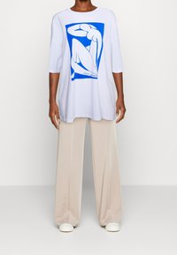 Camiseta blanca con un gráfico de arte lineal azul de una figura; corte holgado, cuello redondo y mangas de tres cuartos, combinada con pantalones beige de pierna ancha.