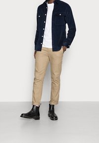 Svarta läderankelstövlar, beige chinos, och en marinblå manchesterklicka över en vit T-shirt, med knappstängning fram och bröstfickor.