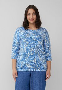 Blaues und cremefarbenes gemustertes Oberteil mit Dreiviertelärmeln, das eine Mischung aus abstrakten Formen und glatter Textur aufweist, kombiniert mit Jeans.