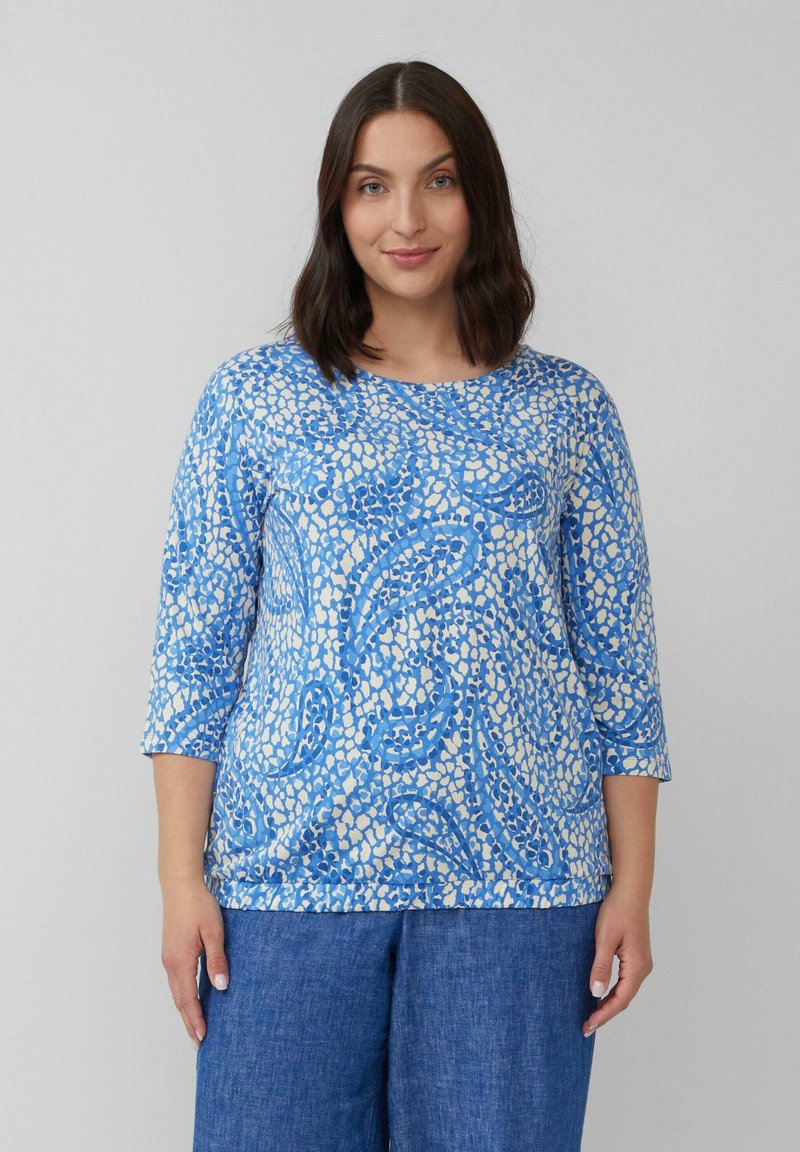Blaues und cremefarbenes gemustertes Oberteil mit Dreiviertelärmeln, das eine Mischung aus abstrakten Formen und glatter Textur aufweist, kombiniert mit Jeans.