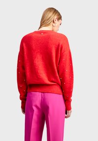 Pull en tricot rouge avec une finition texturée, un col rond et des accents décoratifs en perles le long des manches. Associé à un pantalon rose.