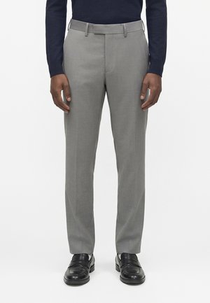 Mand iført skræddersyede lysegrå habitbukser, sorte læderloppersko og en mørk langærmet skjorte, stående foran en ensfarvet baggrund.