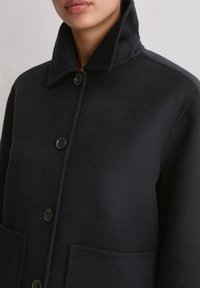 Czarny wełniany płaszcz z wysokim kołnierzem, zapinany z przodu na guziki, z dwoma dużymi kieszeniami, o miękkiej fakturze i wyraźnej sylwetce.