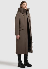 Langer brauner Steppmantel mit hohem Kragen, Reißverschluss vorne und Seitentaschen. Der Stoff wirkt weich und strukturiert und hat eine gerade Passform.