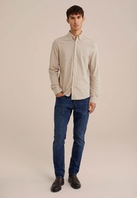 Camisa de manga larga beige con cuello abotonado, de tejido ligero, combinada con jeans ajustados azul oscuro y mocasines marrones. Fondo neutro.