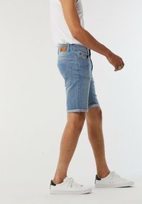Shorts en denim bleu clair avec des ourlets retroussés, cinq poches et une étiquette en cuir brun au dos. Associés à des baskets blanches avec des accents noirs.