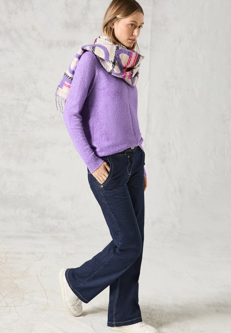 Maglione lavorato a maglia viola, jeans svasati blu scuro e una sciarpa con un motivo dai toni rosa e giallo. La texture appare morbida e accogliente.