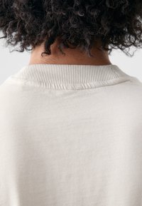 Krämfärgad sweatshirt med ribbad krage och struktur. Bakifrån syns slät tyg och naturliga lockar i nacken.