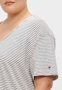Stripet kortermet topp i krem og marineblå, har V-hals og en liten logoakseent på ermet. Mykt, strukturert stoff.