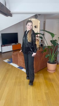 Sort bluse med palietter og lange ærmer, båret med sorte bukser og sorte sko. Inde i et rum med trægulv og en grøn plante i nærheden.