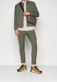 Olivgrön jacka och byxor i en åtsittande stil, beige skjorta under. Svarta och orange sneakers, grå axelväska med dragkedja.