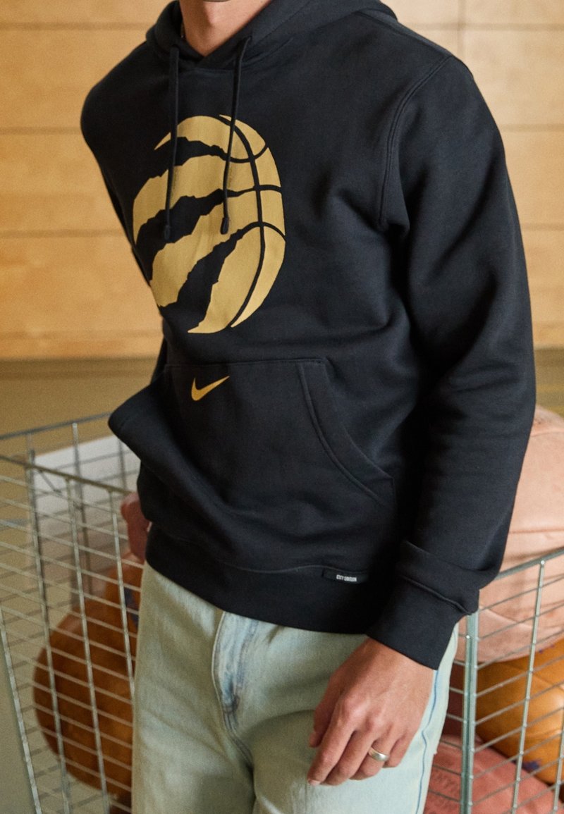 Personne portant un sweat à capuche noir avec un logo doré de basketball et de griffe, debout près d'un panier en fil métallique rempli de ballons de basketball.