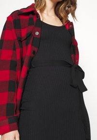 Chemise en flanelle à carreaux rouges et noirs avec des boutons sur une robe noire côtelée ajustée. La robe présente un détail de nœud sur le côté.