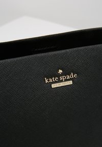 Borsa nera testurizzata con logo dorato "kate spade NEW YORK". Presenta un design elegante e un materiale liscio e resistente.