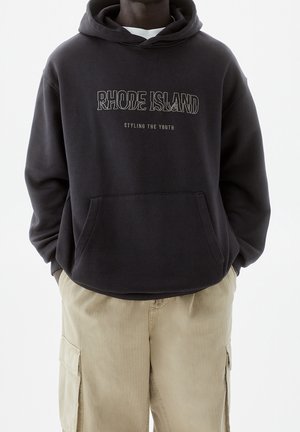 Haut à capuche en coton noir avec poche kangourou à l'avant. Présente le texte "RHODE ISLAND" et "STYLING THE YOUTH" imprimé en blanc et en gris clair.