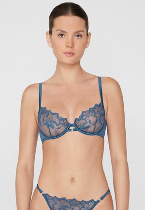 Jeune femme aux cheveux attachés portant un soutien-gorge en dentelle bleue à motifs floraux et une lingerie assortie sur fond uni.