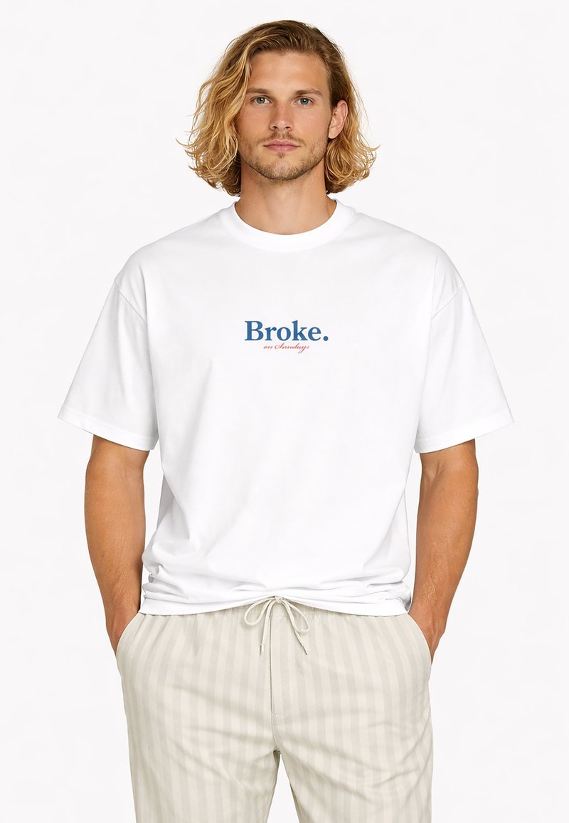 Homme aux cheveux blonds mi-longs portant un T-shirt blanc avec l'inscription « Ruiné. le samedi » et un pantalon à cordon de serrage à rayures claires.