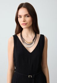 Meerlagige ketting met zwarte koorden, versierd met gouden, zilveren en gestructureerde zilveren metalen accenten. Draagt door een model in een zwarte outfit.