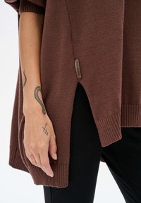 Brązowy sweter dzianinowy o luźnym kroju, z prążkowanymi mankietami i dołem, rozcięciem po bokach oraz metką z napisem "Blue Shadow". Widoczne czarne spodnie.