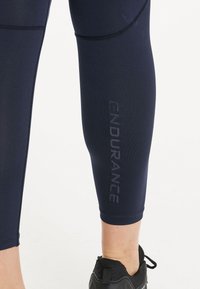 Sotilasnavy urheiluleggingsit, joissa on tiukka muotoilu, tekstuurinen kangas ja "ENDURANCE"-logo painettuna pystysuoraan sääreen. Mustat jalkineet alla.