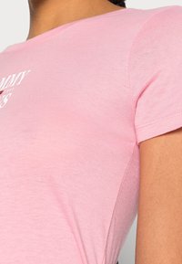 Detailní záběr na osobu v růžovém tričku s krátkým rukávem, na kterém je částečně viditelný bílý a červený text poblíž ramene.